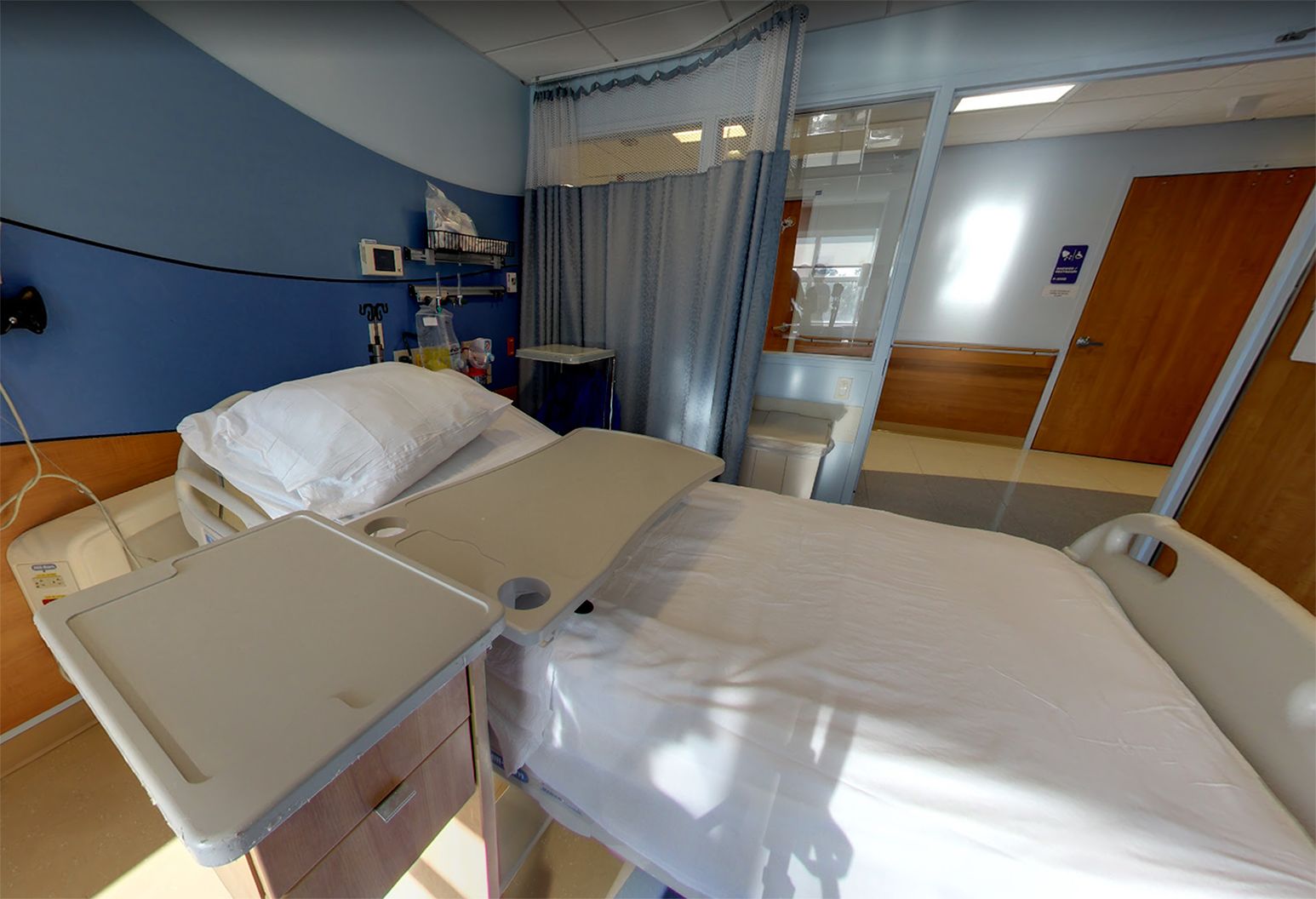 This is a patient room with a sleeper bed, television and a private bathroom with shower. The walls are shades of blue and brown and natural sunlight streams in, evoking a calming and peaceful vibe.